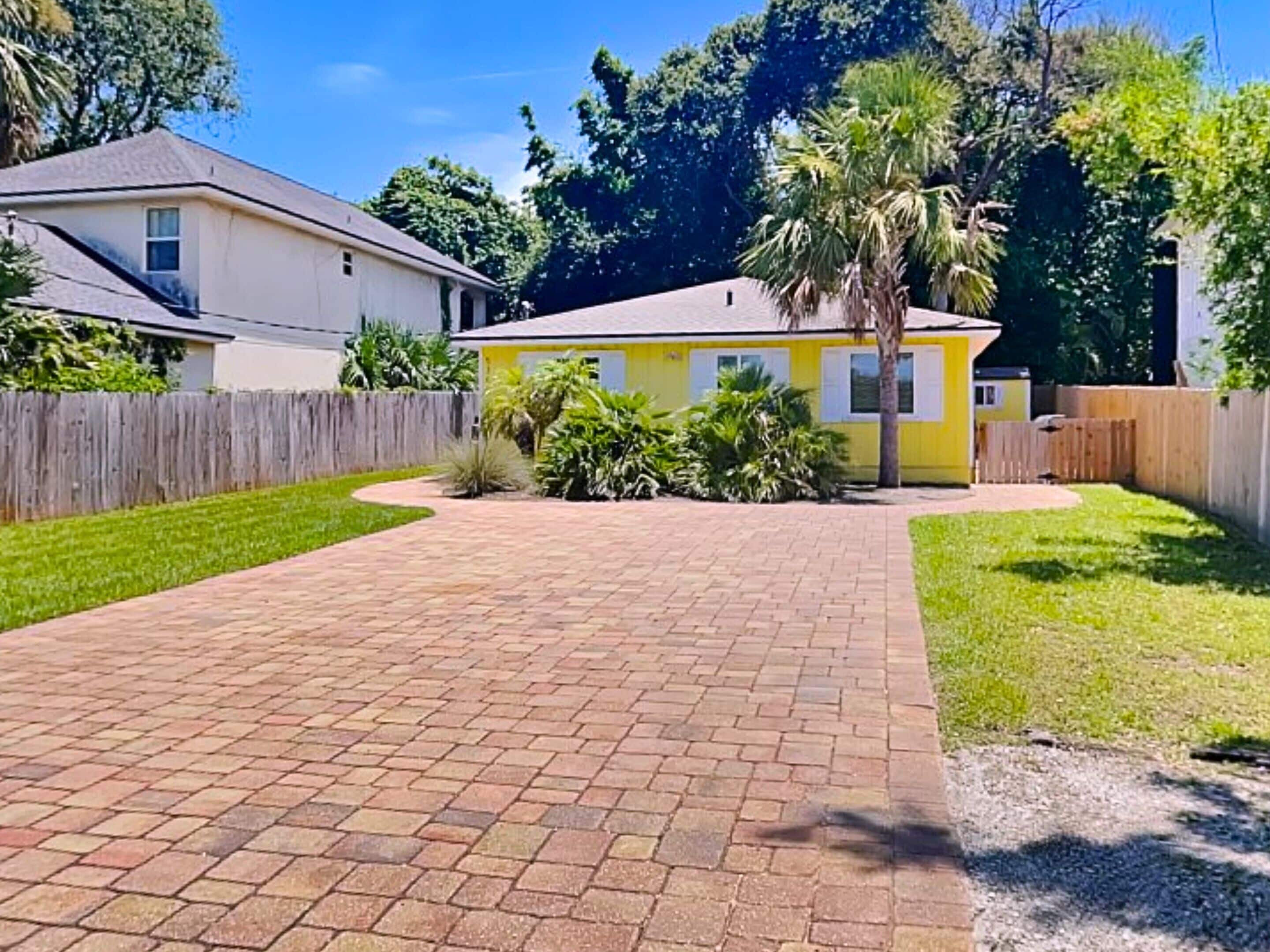 A residential property with a paved driveway, a fenced yard, and a variety of vegetation including palm trees and shrubs.
