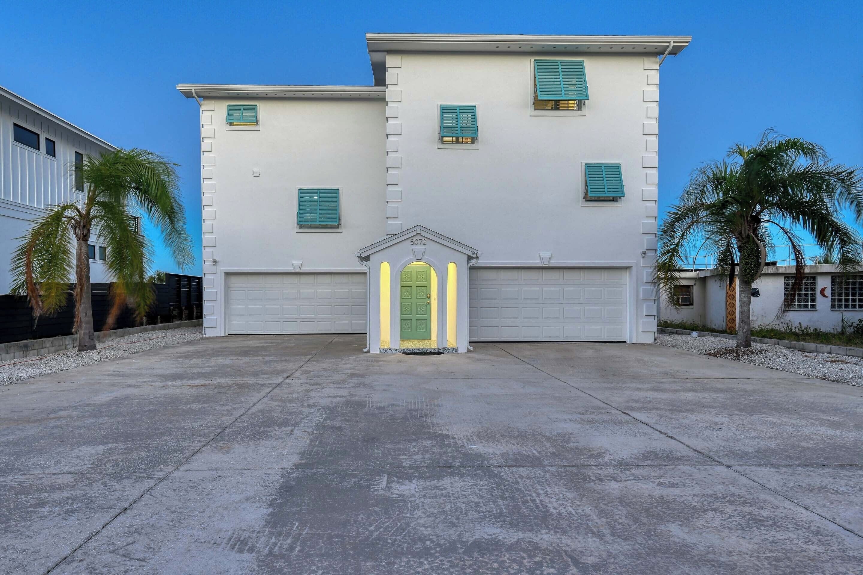 Un edificio de dos pisos con entrada para garage, palmeras y un cielo azul despejado.