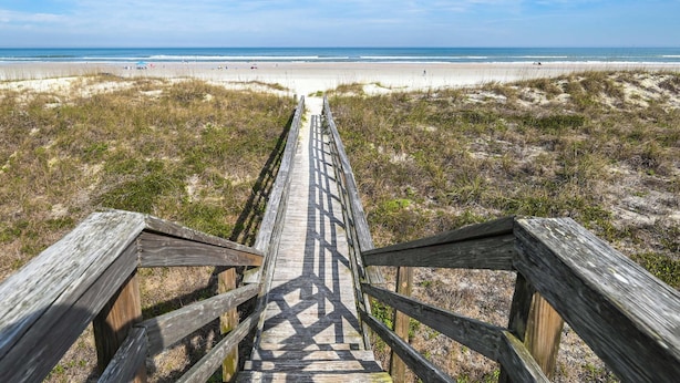 Ein hölzerner Steg führt zu einem Strand mit Dünen und blauem Himmel.
