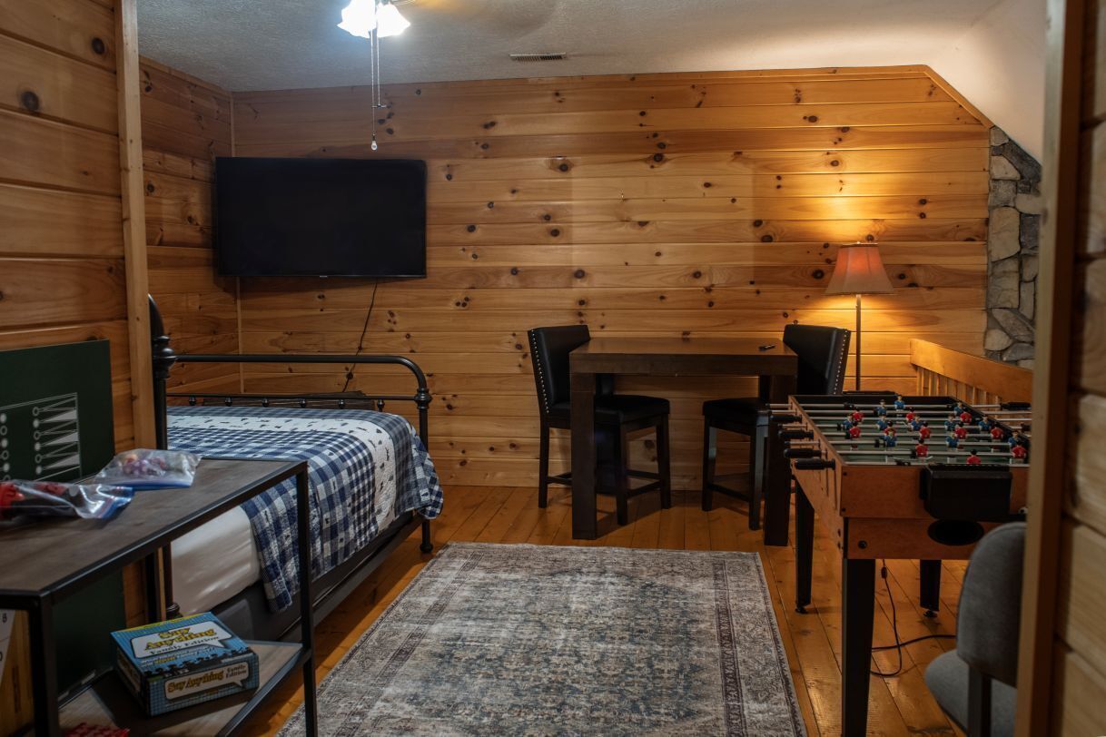 A cozy room with a bed, a table, a foosball table, and a television mounted on the wall.