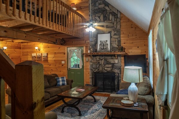 A cozy log cabin interior with a stone fireplace, wooden walls, a green door, and a balcony.