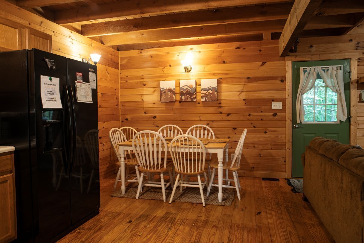 A wooden cabin interior with a dining table, chairs, a refrigerator, and a green door.