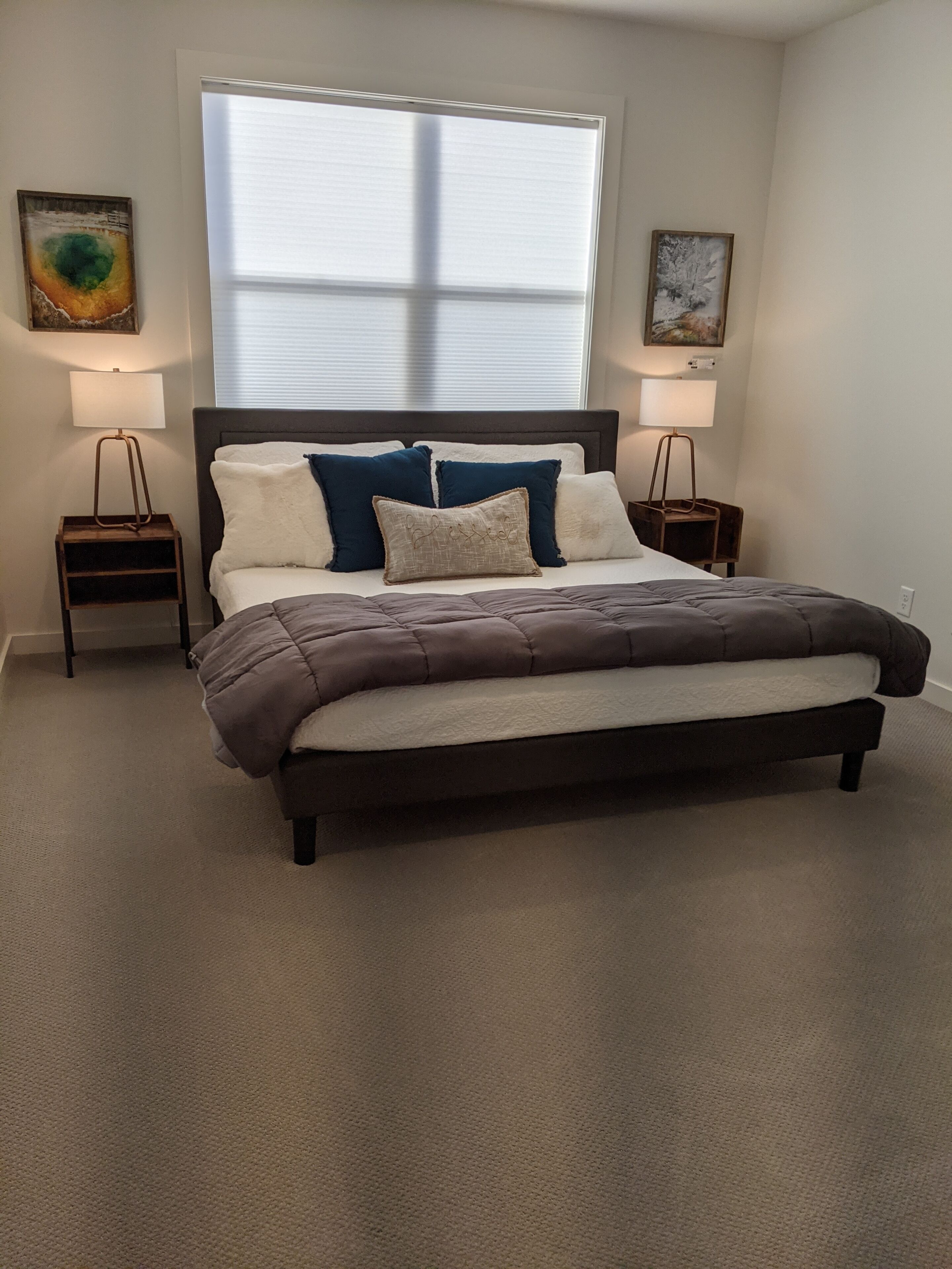 A bedroom with a bed, two bedside tables, a window with blinds, and two wall-mounted lamps.