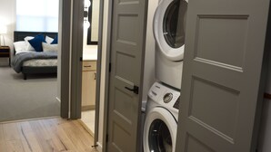 A laundry room with two stacked front-loading washing machines, a grey door, and a glimpse of a bedroom with a bed and a nightstand in the background.
