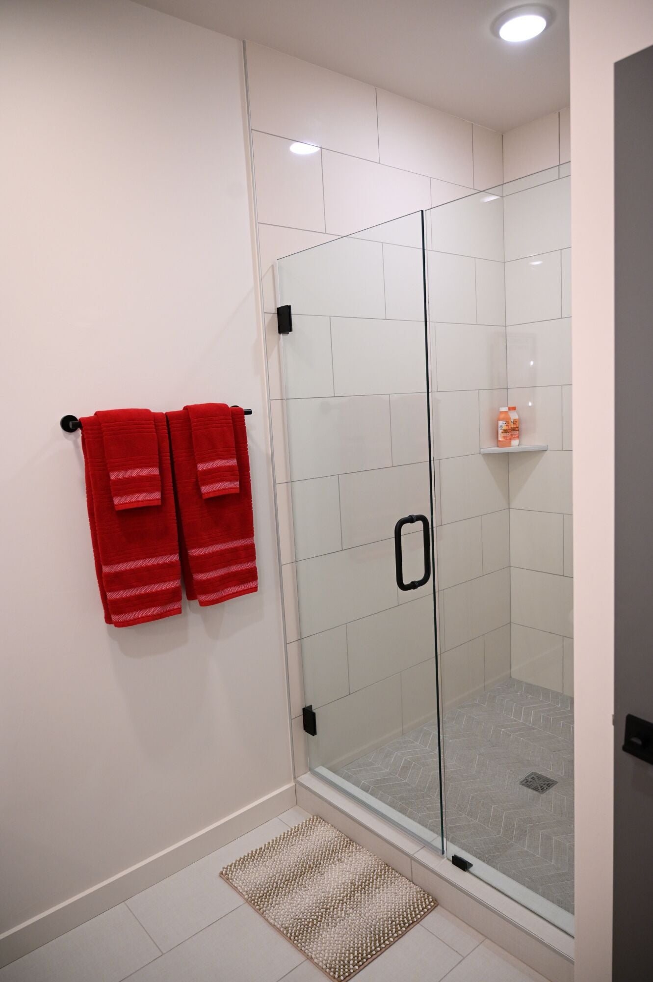 A modern bathroom with a glass shower enclosure, white tiled walls, and a small shelf with toiletries.