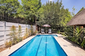 A rectangular swimming pool with clear blue water, surrounded by a tiled deck and a low brick wall. There are two lounge chairs with umbrellas, and a thatched-roof structure to the right. - Retro-vintage New Yorker Apartment in Centurion (Centurion)