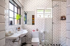 A bathroom with a white sink, toilet, and shower. There are plants on the windowsill and toiletries on the sink. - Retro-vintage New Yorker Apartment in Centurion (Centurion)