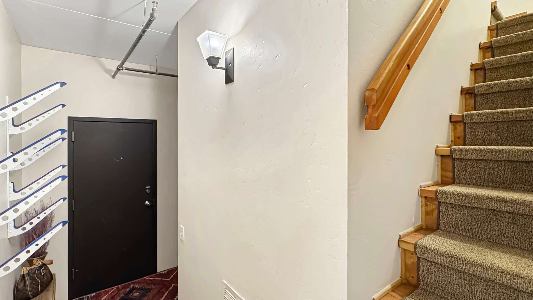 A hallway with a black door, a wall-mounted light fixture, a wooden staircase, and a ceiling-mounted pipe.