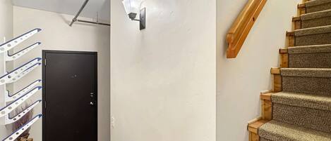 A hallway with a black door, a wall-mounted light fixture, a wooden staircase, and a ceiling-mounted pipe.