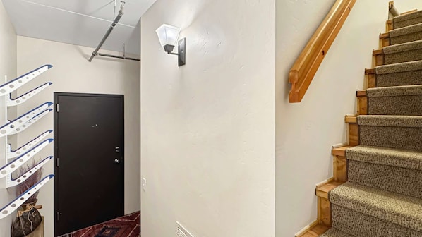 Un pasillo con una puerta negra, una lámpara de pared, una escalera de madera y un tubo en el techo.
