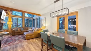 A modern living room with a large sofa, dining table, and chairs, large windows, and a fireplace.