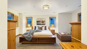 Un dormitorio con una cama grande, cabecera de madera, dos mesitas de noche, un banco y un cuadro en la pared.