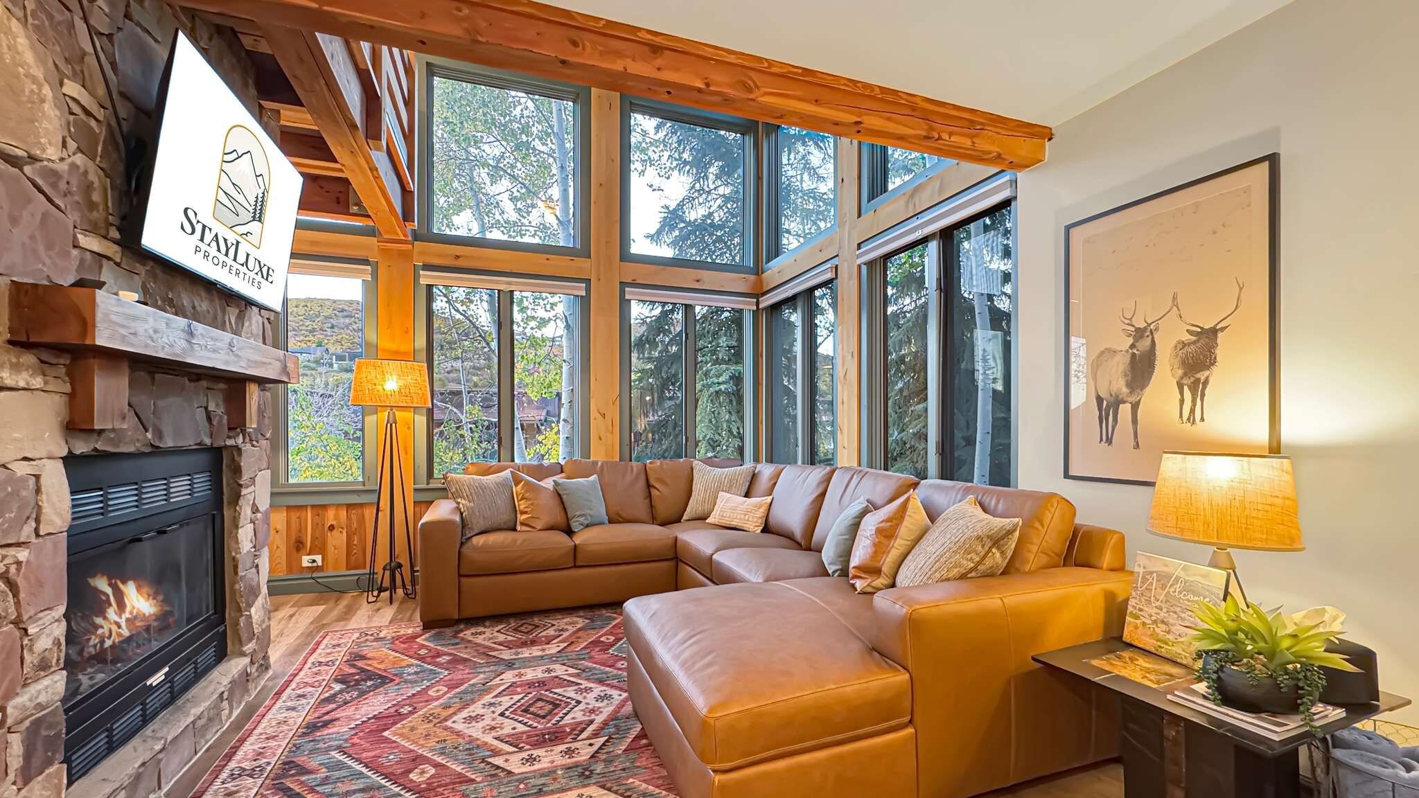 A cozy living room with a stone fireplace, a large window with a view of trees, a leather sofa, and a framed deer artwork.