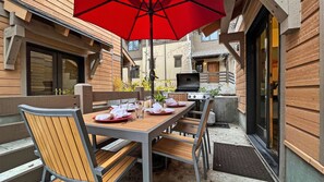 An outdoor dining area with a table set for four, a red umbrella, and a barbecue grill.