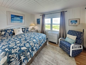A bedroom with a bed, a chair, a nightstand, a window with curtains, and a view of the outdoors.