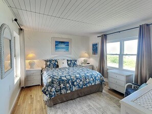 A bedroom with a large bed, two bedside tables, a window with curtains, and a view of greenery outside. - Hunter's Haven by Distinctive Beach Rentals (St. Augustine)