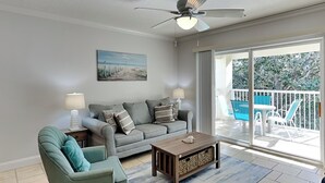Un salon avec un canapé gris, un fauteuil turquoise, une table basse en bois et un ventilateur de plafond. Il y a un tableau au mur et un balcon avec des meubles d’extérieur.