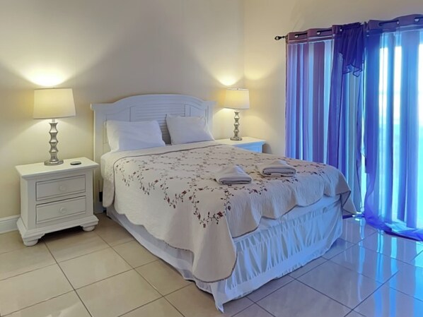 A neatly made bed with a floral bedspread and two pillows, flanked by white nightstands with lamps, in a room with a large window covered by blue curtains. - Wave Runner by Distinctive Beach Rentals (Flagler Beach)