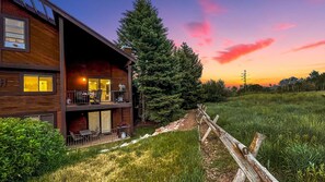 A two-story wooden cabin with a balcony, surrounded by a grassy landscape and a tall evergreen tree.