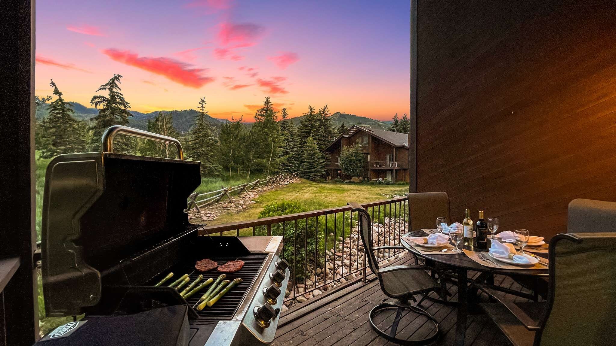 A barbecue grill with steaks and asparagus on a balcony overlooking a mountainous landscape.
