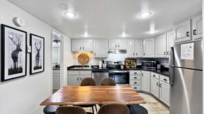 Une cuisine moderne avec des meubles blancs, des appareils en acier inoxydable, une table à manger en bois et deux tableaux représentant des cerfs, encadrés et accrochés au mur.