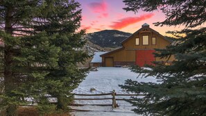 Un granero de madera con puerta roja, rodeado de nieve y árboles de hoja perenne.
