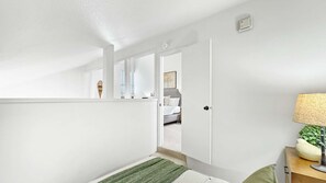 Ein modernes Schlafzimmer mit Bett, Nachttisch, Lampe und Blick auf einen anderen Raum durch eine offene Tür.