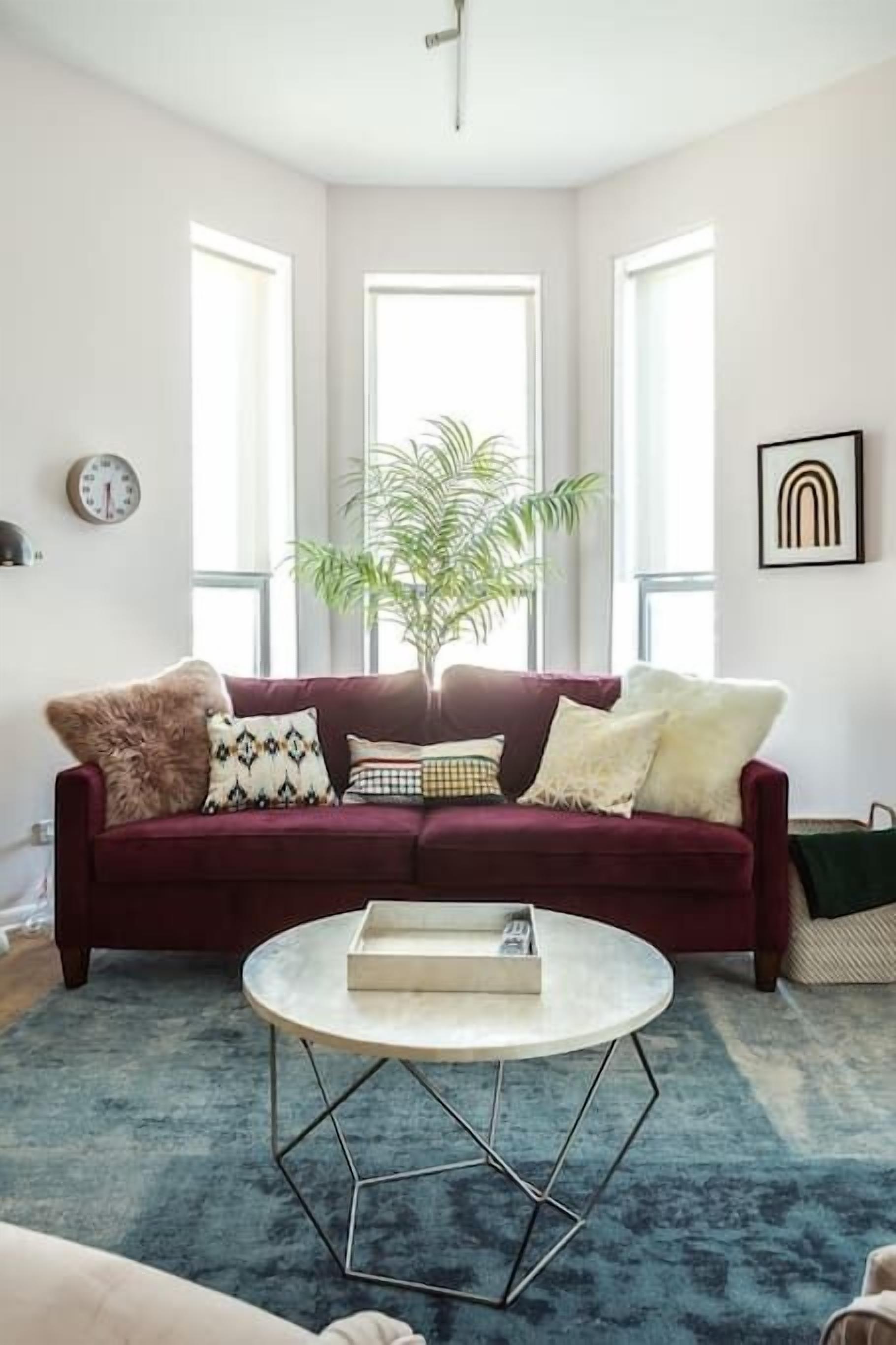 Un salon avec un canapé rouge foncé, une table basse ronde et une grande fenêtre avec une plante.