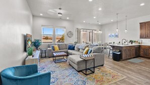 A spacious living room with a sectional sofa, a coffee table, and a blue armchair. A kitchen with wooden cabinets and a countertop is visible in the background. - Ocotillo Springs Resort 69: 3bd, 3.5 bath with a Private Hot Tub, Fire Pit, and Private Patio (Santa Clara)