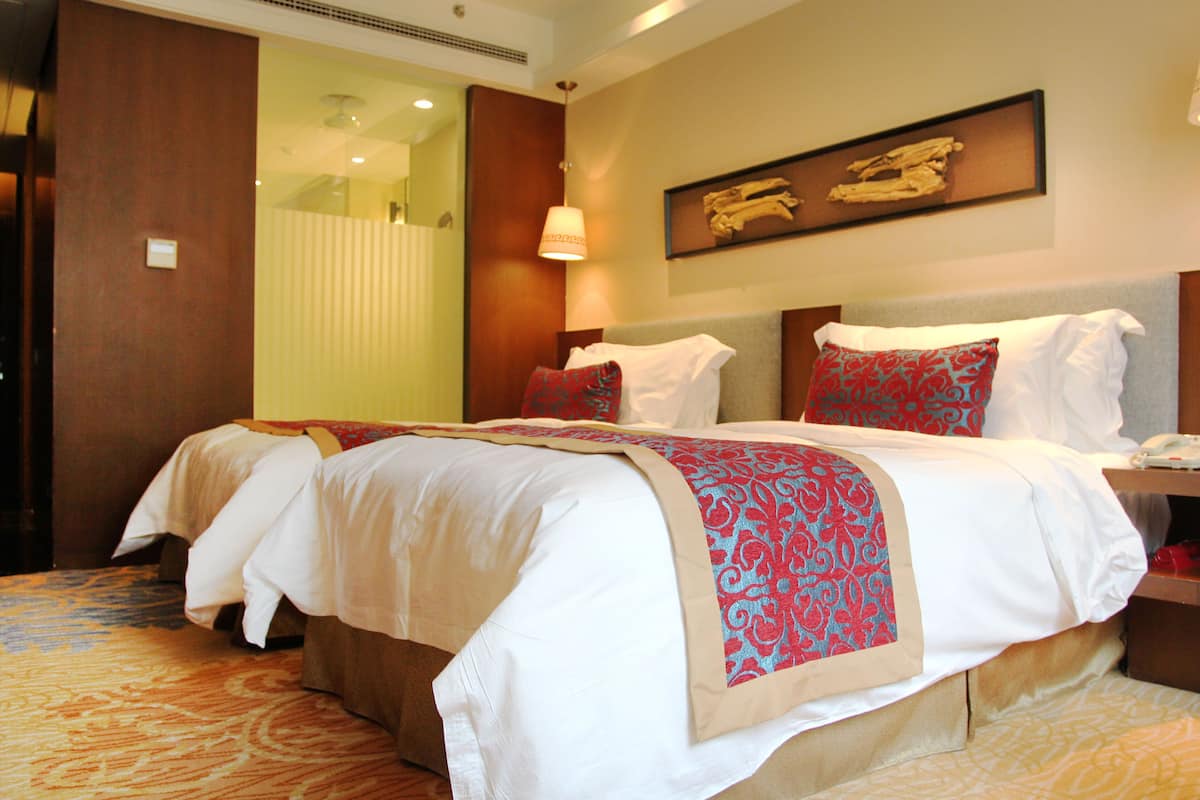 A hotel room with two beds, a large window, a wooden headboard, and a framed artwork on the wall.