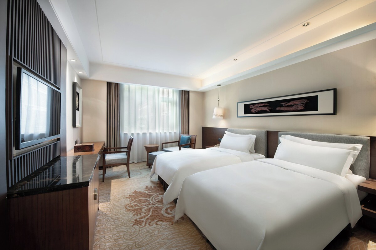 A hotel room with two beds, a desk, a chair, a TV, and a large window with curtains.
