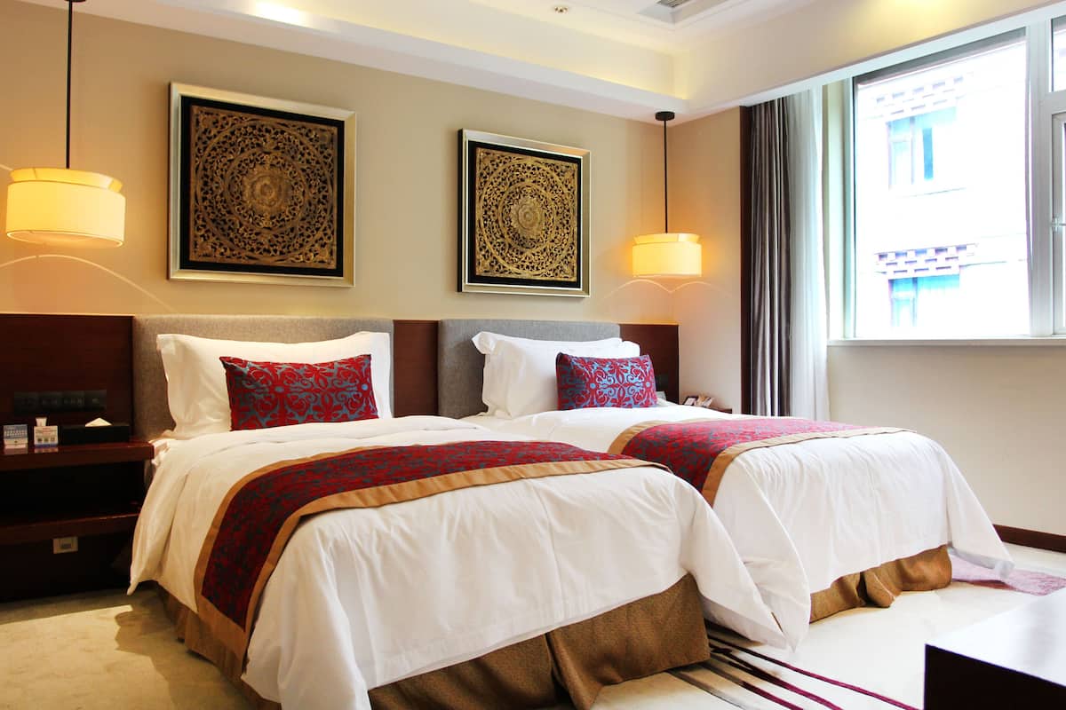 A hotel room with two beds, each with white linens and red and brown decorative pillows. There are two wall-mounted lamps, and the room has a large window with curtains.