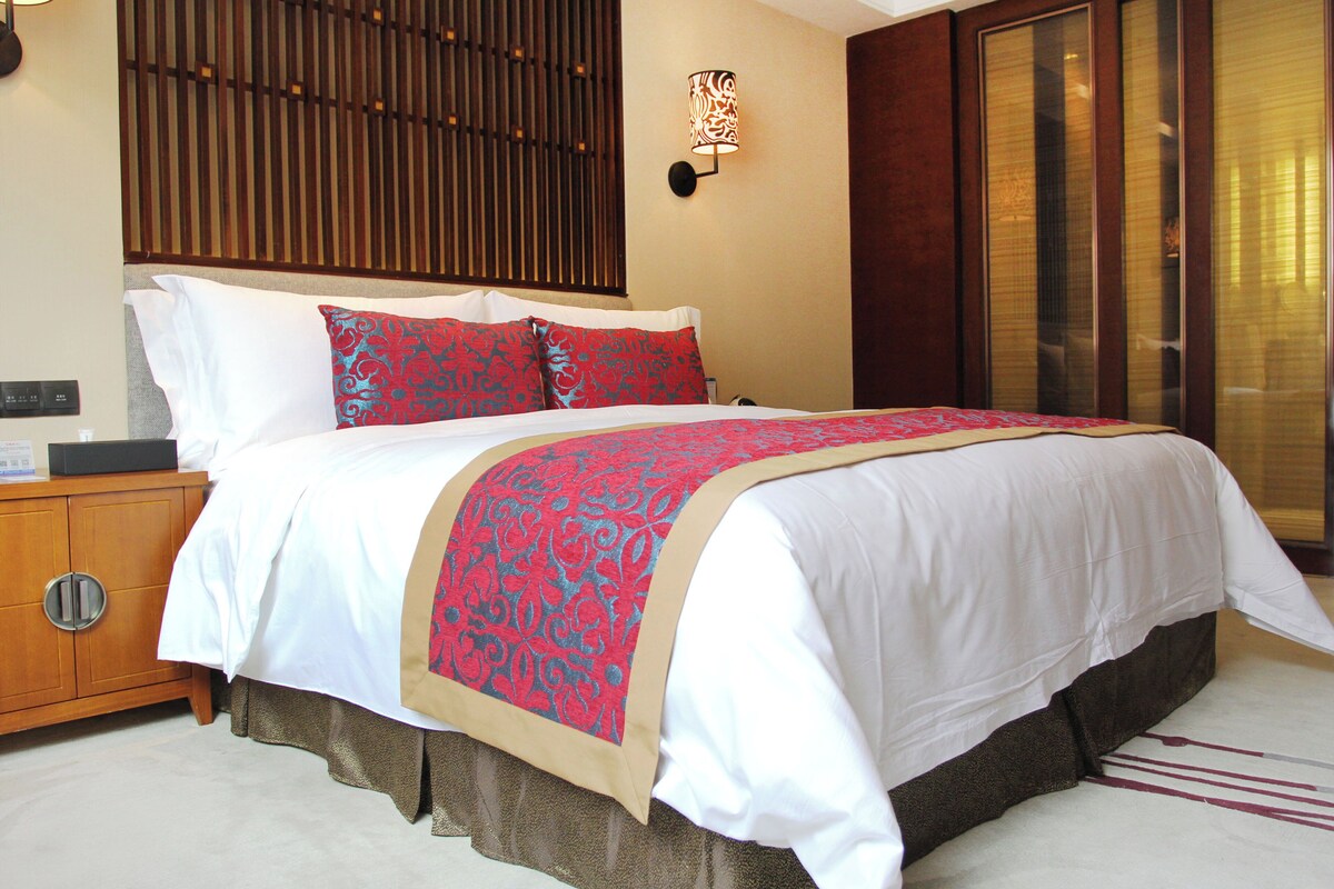 A hotel room with a bed, bedside table, lamp, and a view of the outside.