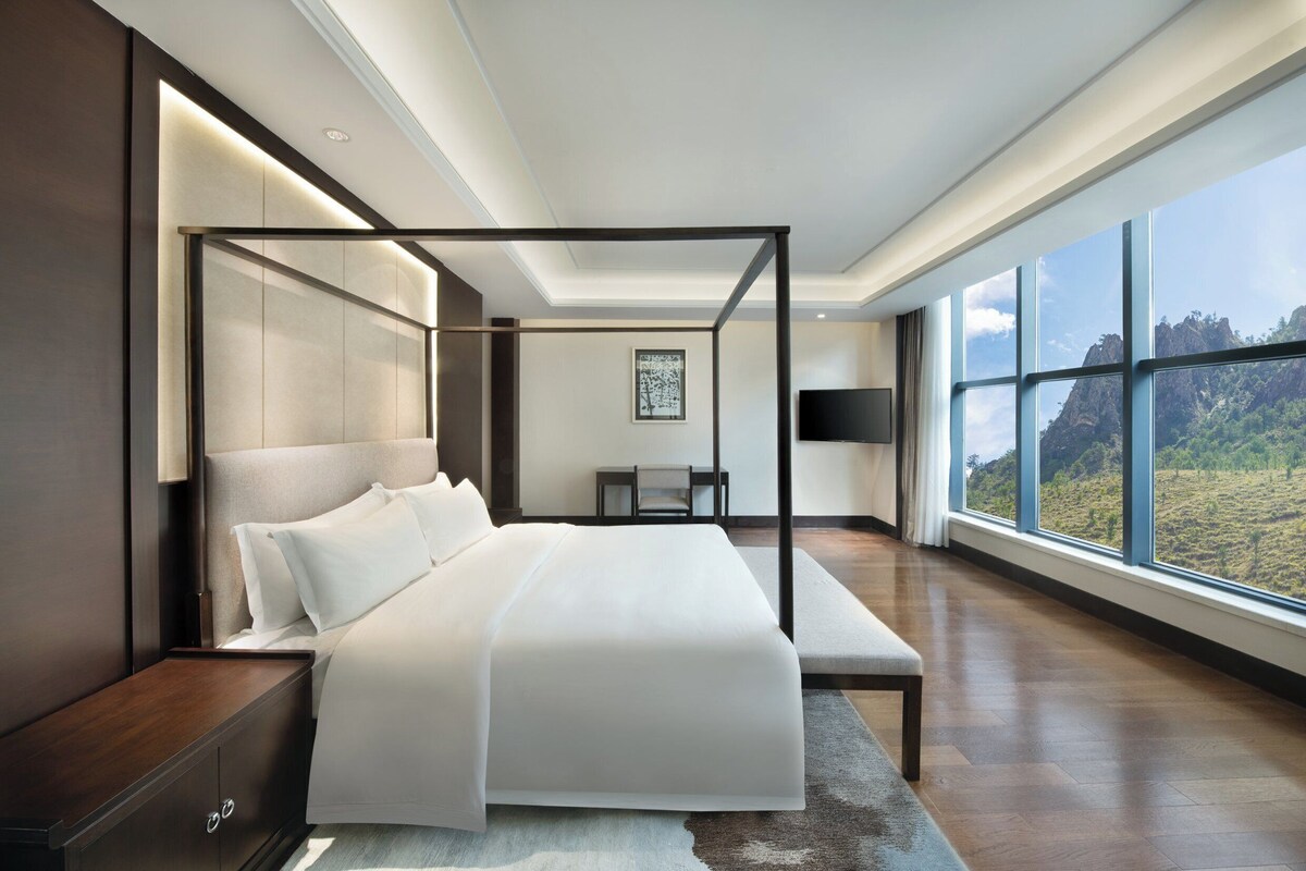 A modern bedroom with a four-poster bed, a large window offering a view of the outdoors, and a minimalist design.