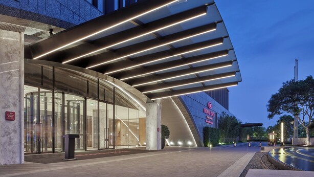 Une entrée d’hôtel moderne, dotée d’une façade en verre incurvé, d’un auvent imposant agrémenté de luminaires linéaires et d’une enseigne bien visible.