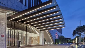 Una entrada de hotel moderna con fachada de vidrio curvo, un pronunciado alero con luces lineales y un letrero visible del hotel.