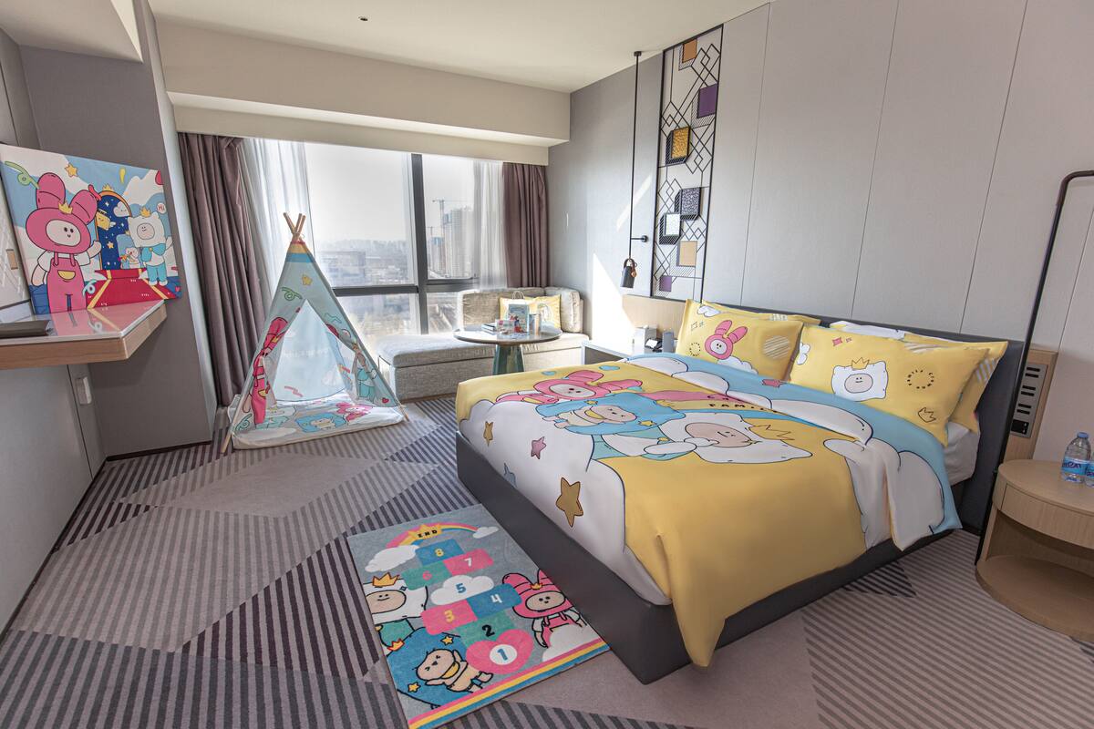 Une chambre d’hôtel avec un grand lit, une fenêtre avec des rideaux, une petite table et un tapis coloré.