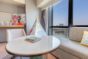 A hotel room with a round table, a sofa, a window with a city view, and a children's play area.