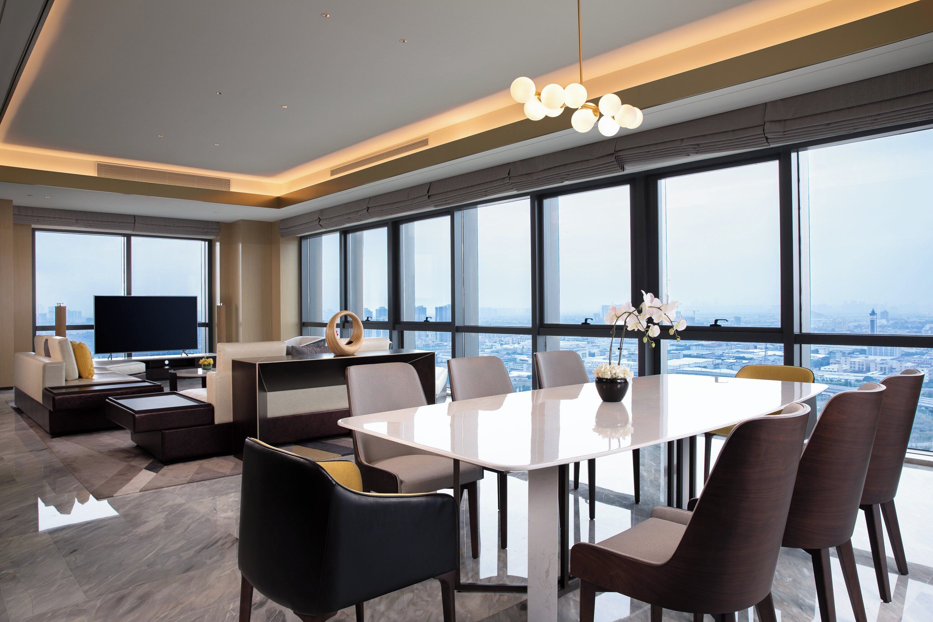 A modern hotel room with a large window, a flat-screen TV, a dining area, and a sofa.