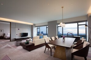 Ein modernes Hotelzimmer mit einem großen Esstisch, bequemen Sitzgelegenheiten, einem Flachbildfernseher und Blick auf die Stadt durch große Fenster.