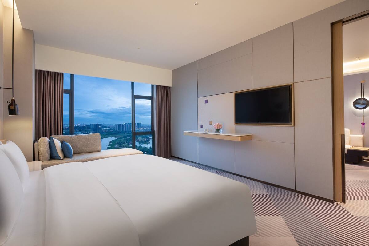 Une chambre d’hôtel moderne avec un grand lit, un téléviseur fixé au mur, une vue sur la ville et une petite table agrémentée d’un vase de fleurs.