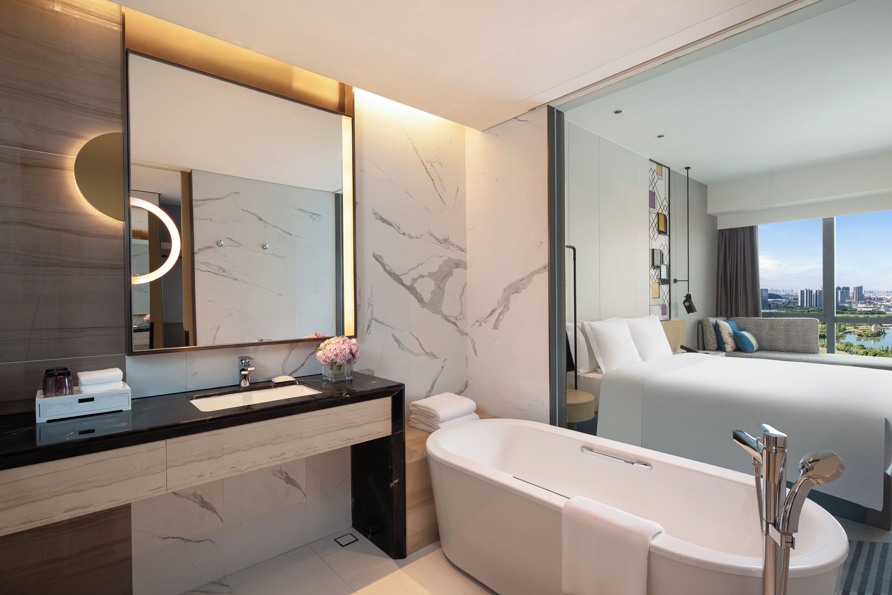 Une salle de bain d’hôtel moderne avec un meuble-lavabo en marbre, un grand miroir, une baignoire et une vue sur la ville par la fenêtre.