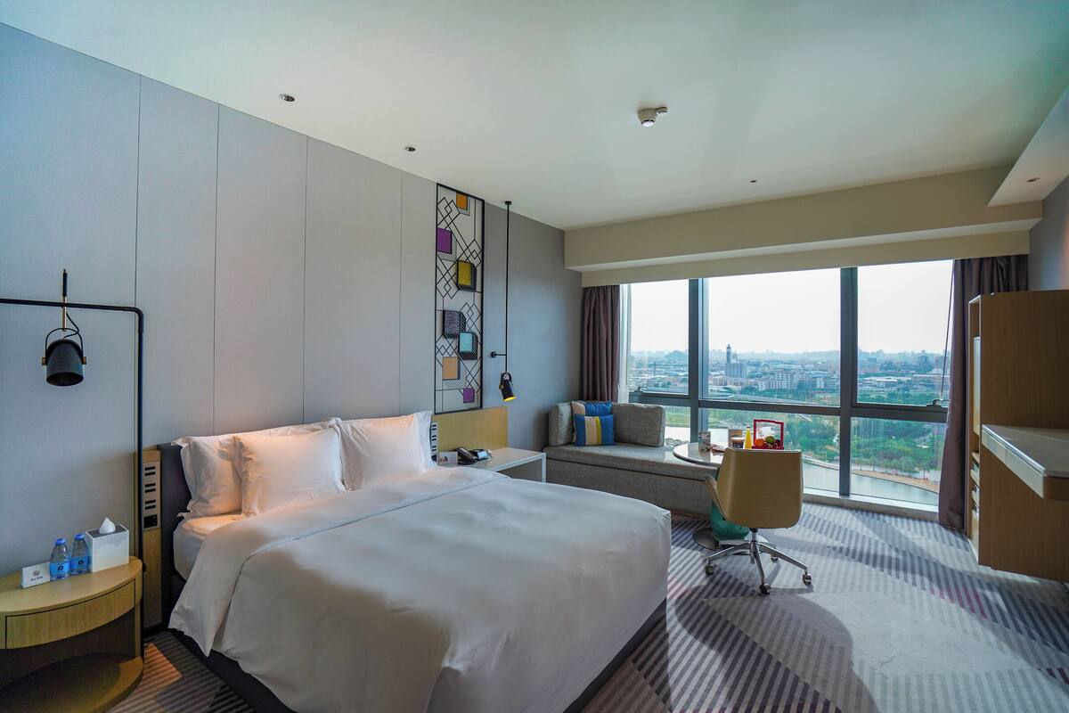 Une chambre d’hôtel dotée d’un grand lit, d’une table de chevet avec des commodités, d’une fenêtre donnant sur la ville et d’un coin salon avec vue.