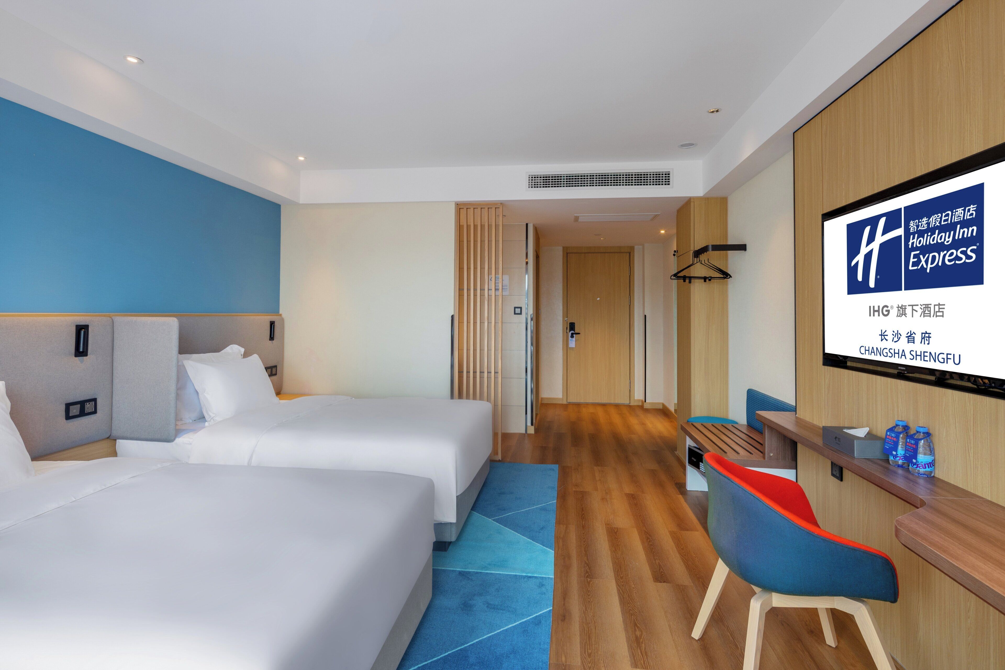A hotel room with two beds, a desk, a chair, and a large screen displaying the hotel name.
