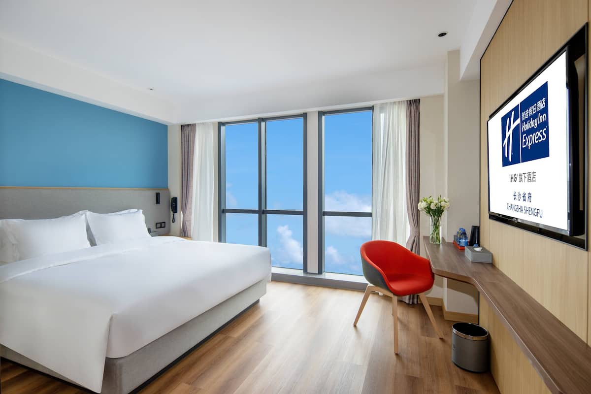 A hotel room with a large bed, a desk with a chair, a window with a view of the sky, and a screen displaying hotel information.