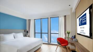 A hotel room with a large bed, a desk with a chair, a window with a view of the sky, and a screen displaying hotel information.