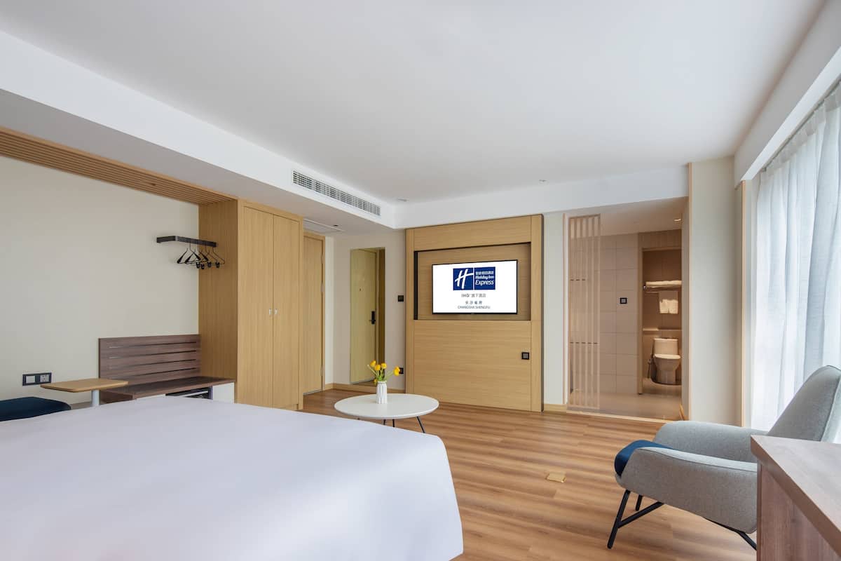 A modern hotel room with a large bed, a seating area with a table and chairs, a television mounted on the wall, and a view of the bathroom and hallway.