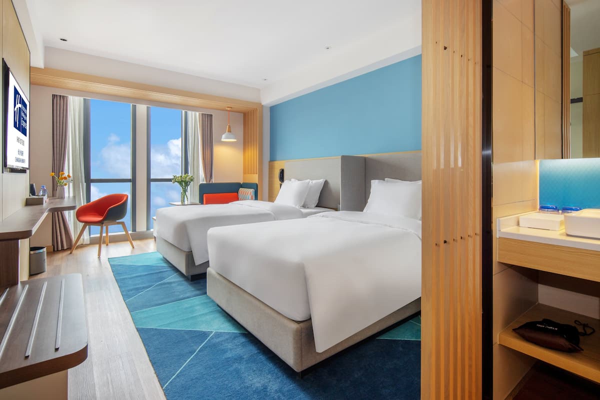 A hotel room with two beds, a desk, a chair, and a large window offering a view.