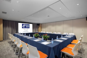 Une salle de conférence avec une longue table, des chaises blanches dotées de coussins orange, des bouteilles d’eau et un écran affichant du texte.