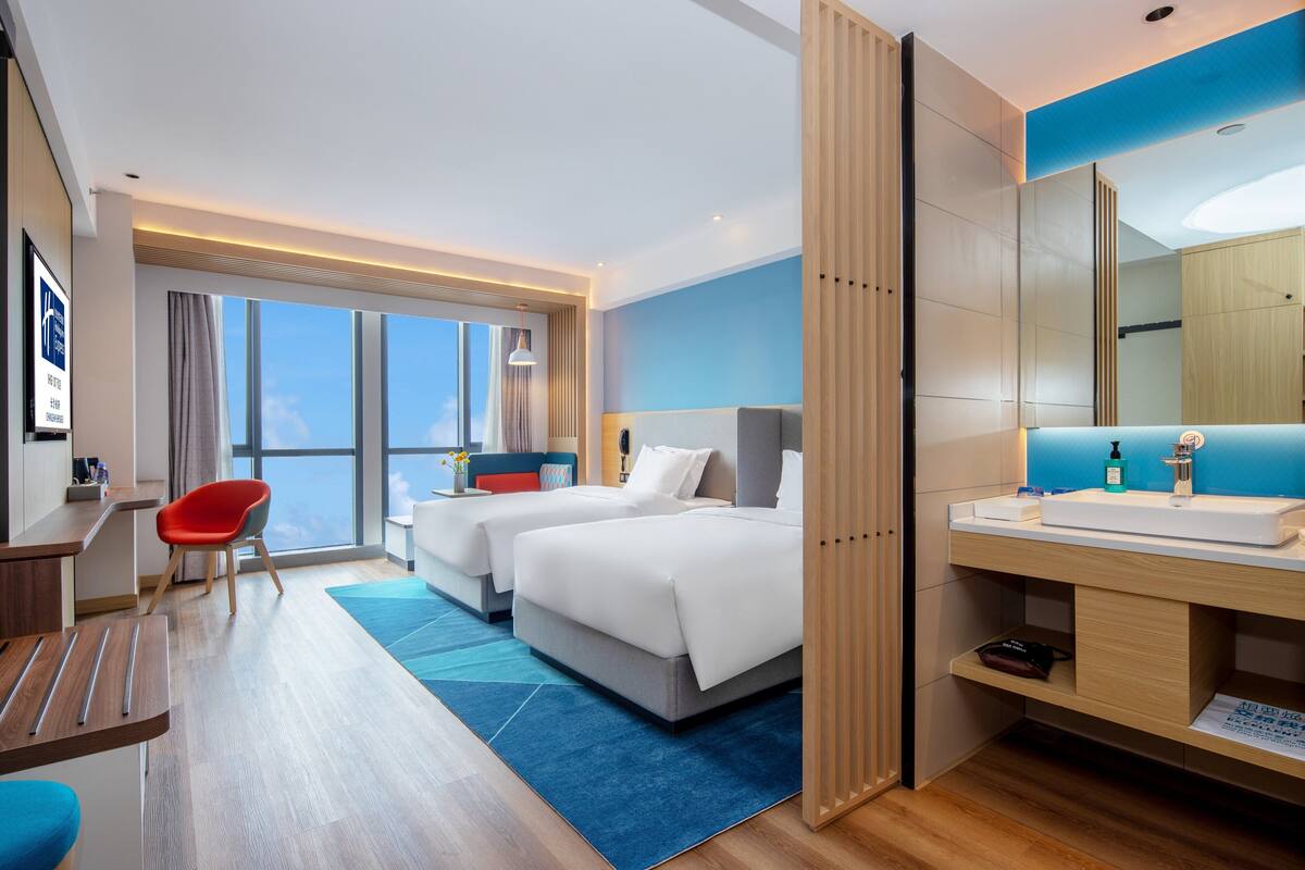 A hotel room with two beds, a desk, a chair, a large window, and a bathroom with a sink and mirror.